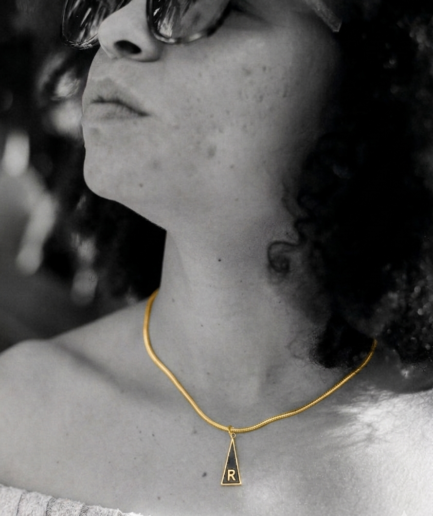 Imagen de estilo lifestyle que muestra la caída y el tamaño de la Medalla Inicial "S" - Serie Triangular. Se aprecia el contraste del esmalte negro con el relieve en oro de 18K sobre la piel.